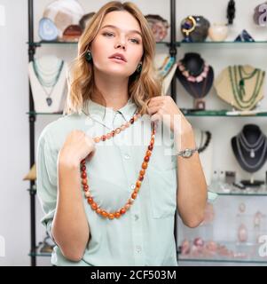 Jeune femme élégante cliente choisissant des bijoux en pierres précieuses naturelles dans la boutique, essayant sur des perles de sardonyx orange poli Banque D'Images