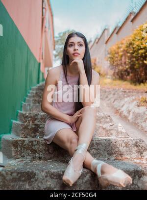 Jeune brune ballerine à pointe chaussures assis sur des marches altérées et tout en vous reposant après l'entraînement contre le mur rayé dans la rue Banque D'Images