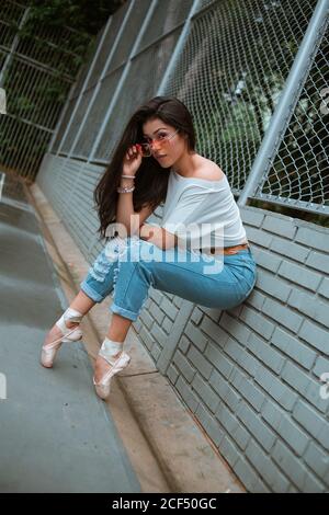 Vue latérale de la ballerine femme mince dans des vêtements décontractés et des lunettes de soleil assis sur le pied à la pointe avec des chaussures de pointe sur la barrière murale en brique sur la rue Banque D'Images