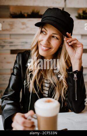 Belle femme blonde à cheveux longs et tendance, buvant un verre de délicieux café mousseux tout en regardant loin Banque D'Images