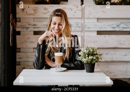 Belle femme blonde à cheveux longs et tendance, buvant un verre de délicieux café moussant avec les yeux fermés Banque D'Images