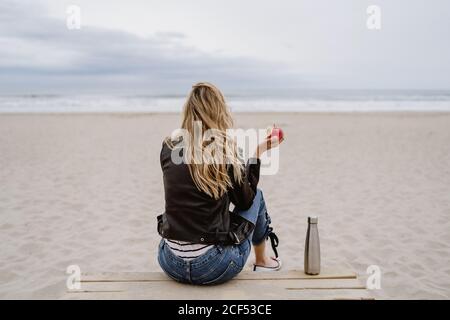 Vue arrière de la femme blonde tendance non reconnaissable en casquette noire et veste en cuir avec pomme mûre rouge à la main sur la plage de sable Banque D'Images