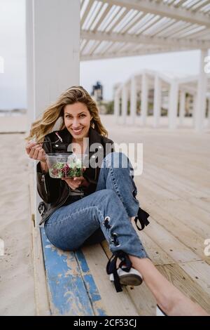 Élégante femme blonde aux cheveux longs en blouson noir appréciant une salade verte saine tout en étant assise sur une terrasse en bois sur la côte en souriant à l'appareil photo Banque D'Images