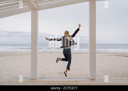 Vue arrière de la femme tendance et méconnaissable en blouson noir sautant avec les bras levés sur une jetée en bois blanc avec l'océan en arrière-plan Banque D'Images