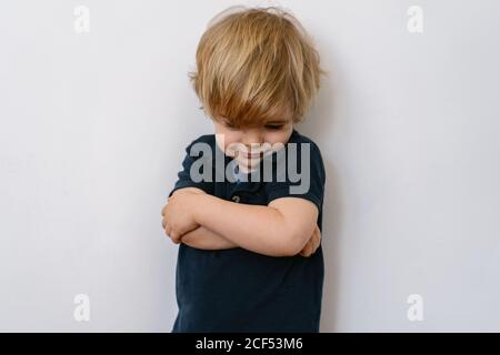 Un petit garçon blond agacé dans des vêtements décontractés qui regardent vers le bas insatisfaction lorsqu'on se tient debout sur un mur blanc avec des bras croisés Banque D'Images