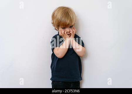 Un petit garçon blond agacé dans des vêtements décontractés qui regardent loin avec insatisfaction lorsqu'on se tient debout sur un mur blanc avec les mains sur le visage Banque D'Images