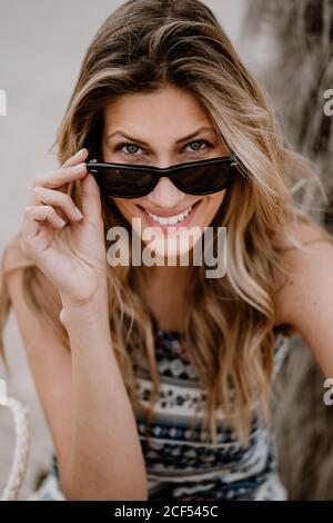 Portrait en gros plan d'une jolie femme blonde en lunettes de soleil noires, assise sur le sable et regardant l'appareil photo Banque D'Images