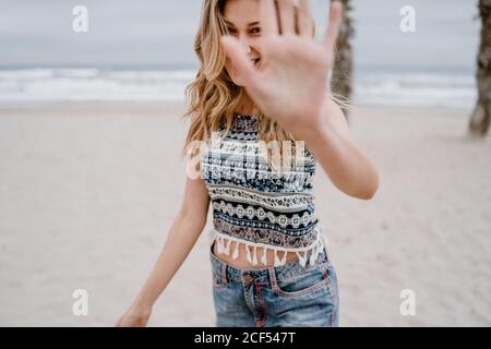 Blonde gaie femme en haut coloré et short jean lui bloquant le visage avec la main sur la mer Banque D'Images