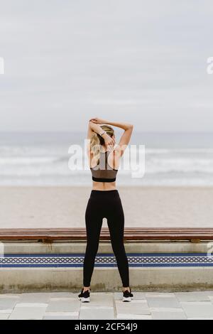 Vue arrière d'une athlète féminine anonyme en tenue active debout avec les mains levées en position verrouillée et s'étirant près du bord de mer par temps nuageux Banque D'Images