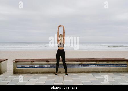 Vue arrière d'une athlète féminine anonyme en tenue active debout avec les mains levées en position verrouillée et s'étirant près du bord de mer par temps nuageux Banque D'Images