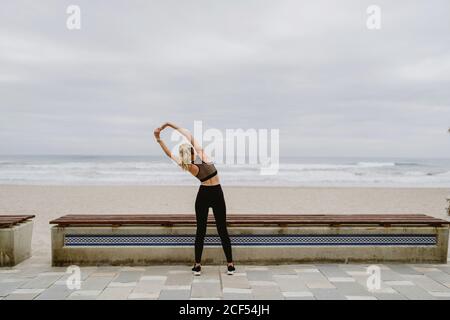 Vue arrière d'une athlète féminine anonyme en tenue active debout avec les mains levées en position verrouillée et s'étirant près du bord de mer par temps nuageux Banque D'Images