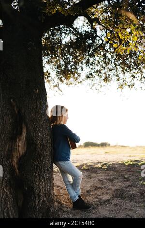 Adorable petite fille sérieuse en tenue décontractée jouant ukulele guitare tout en se tenant près de l'arbre dans la journée ensoleillée d'été dans la campagne Banque D'Images