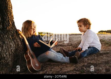 Vue latérale d'une adorable petite fille lisant une histoire intéressante à frère plus jeune assis sous l'arbre avec une guitare ukulele en été dans la campagne Banque D'Images