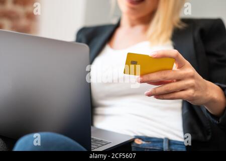 Courte femme blonde non reconnaissable dans des vêtements décontractés entrant dans la carte de crédit des références sur un ordinateur portable tout en étant assis dans un fauteuil confortable et en faisant achats en ligne à la maison Banque D'Images