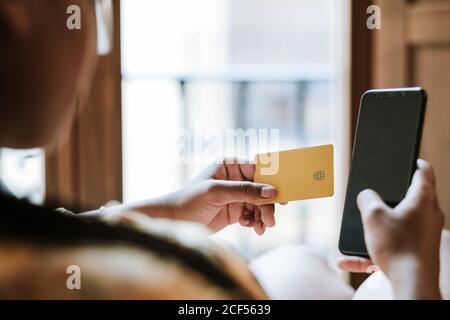 Vue arrière de Crop anonyme ethnique femelle avec carte de crédit et un smartphone qui effectue des achats en ligne à la maison Banque D'Images