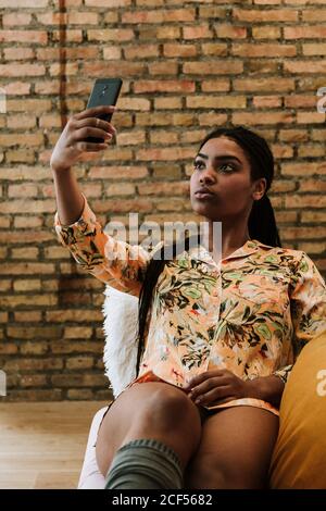 Jeune femme afro-américaine sérieuse tenue décontractée prenant le selfie avec un smartphone assis sur une chaise contre un mur de briques appartement moderne Banque D'Images