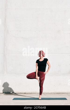Athlète féminine confiante et assiégée dans les vêtements de sport méditant en se tenant debout position vrikshasana avec les mains fermées à la poitrine dans le namaste sur l'entraînement de tapis de sport seul sur la rue contre le mur de béton en plein soleil Banque D'Images