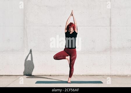 Athlète féminine confiante et assiégée dans les vêtements de sport méditant en se tenant debout position vrikshasana avec les bras relevés et le namaste sur le tapis de sport entraînement seul dans la rue contre le mur de béton à la lumière du soleil Banque D'Images