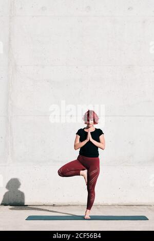 Athlète féminine confiante et assiégée dans les vêtements de sport méditant en se tenant debout position vrikshasana avec les mains fermées à la poitrine dans le namaste sur l'entraînement de tapis de sport seul sur la rue contre le mur de béton en plein soleil Banque D'Images