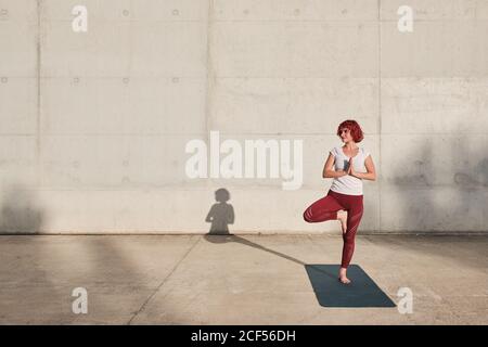 Athlète féminine confiante et assiégée dans les vêtements de sport méditant en se tenant debout position vrikshasana avec les mains fermées à la poitrine dans le namaste sur l'entraînement de tapis de sport seul sur la rue contre le mur de béton en plein soleil Banque D'Images