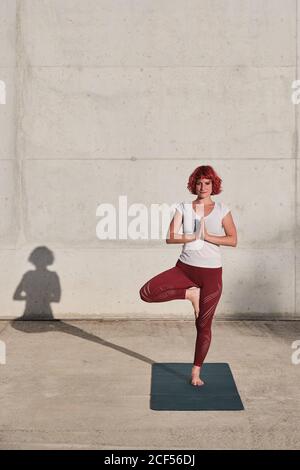 Athlète féminine confiante et assiégée dans les vêtements de sport méditant en se tenant debout position vrikshasana avec les mains fermées à la poitrine dans le namaste sur l'entraînement de tapis de sport seul sur la rue contre le mur de béton en plein soleil Banque D'Images