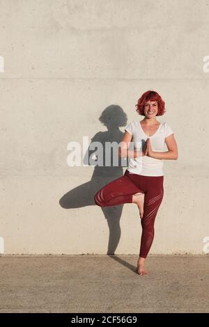 Athlète féminine confiante et assiégée dans les vêtements de sport méditant en se tenant debout position vrikshasana avec les mains fermées à la poitrine dans le namaste sur l'entraînement de tapis de sport seul sur la rue contre le mur de béton en plein soleil Banque D'Images
