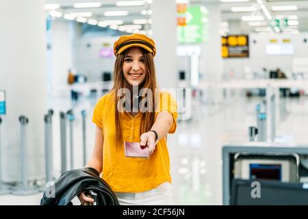 Femme souriante et gaie dans des vêtements et écouteurs élégants et colorés passeport et regarder l'appareil photo lors de l'enregistrement comptoir dans le terminal de l'aéroport Banque D'Images