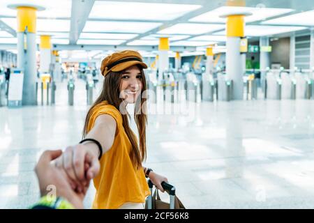 Jeune femme souriante dans un ensemble élégant dirigeant quelqu'un à la main et regarder la caméra sur le chemin de la salle d'attente avec valise dans le terminal de l'aéroport Banque D'Images