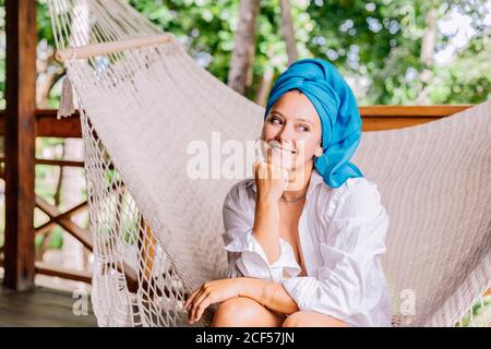 Femme heureuse en chemise et tête de châle lumineuse assis dans un hamac regardant loin par des arbres verdoyants au Costa Rica Banque D'Images