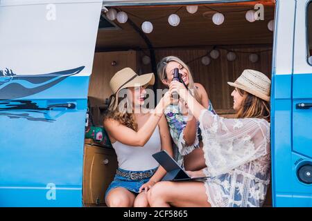 Souriant gai dames avec de longs cheveux dans le chapeau levant des bouteilles de boisson et de clinking pour les vacances en voiture Banque D'Images