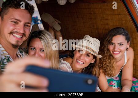 Groupe agréable de jeunes allongé sur le tronc de lumineux minivan et s'amuser à prendre le selfie sur un téléphone mobile plage en journée ensoleillée Banque D'Images