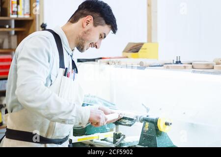 un jeune homme aux cheveux foncés et aux lunettes tient une tige dans ses mains et traite un produit en bois sur un tourner dans l'atelier Banque D'Images