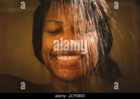 Belle femme ethnique souriant gaiement et gardant les yeux fermés tout en se lavant dans la douche à la maison Banque D'Images