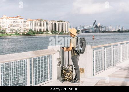 Jeune homme avec une valise et un sac à dos, arrivant sur les rives de Miami, regardant la rivière et les bâtiments un matin ensoleillé Banque D'Images