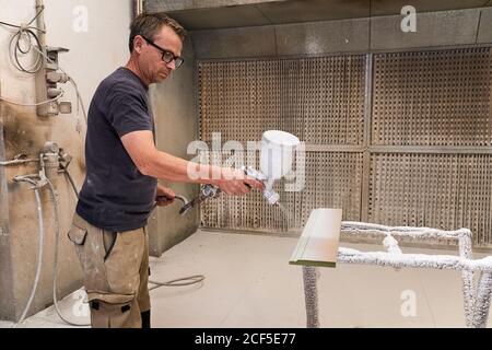 Vue latérale d'un charpentier d'âge moyen sérieux à l'aide d'un pistolet arrosoir pour peindre les détails du bois avec un revêtement ignifuge mousse blanc travailler dans une salle de travail moderne et légère Banque D'Images