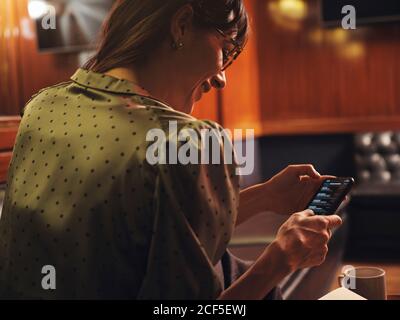 Vue latérale d'une femme élégante et attentionnés surfant sur un téléphone portable confortable assis au canapé en cuir noir dans le café Banque D'Images