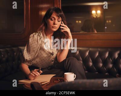 Femme élégante écoutant attentivement les informations sur le téléphone portable et écrivant dans un ordinateur portable confortablement assis à une table ronde dans un café Banque D'Images