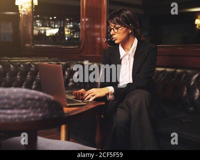 Femme d'affaires intelligente en lunettes dactylographiant sur ordinateur portable confortablement assis dans un canapé en cuir noir dans le café Banque D'Images