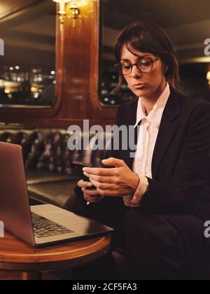 Femme d'affaires intelligente en lunettes dactylographiant sur un ordinateur portable et vérifiant les informations dans un téléphone mobile confortablement assis dans un canapé en cuir noir dans un café Banque D'Images