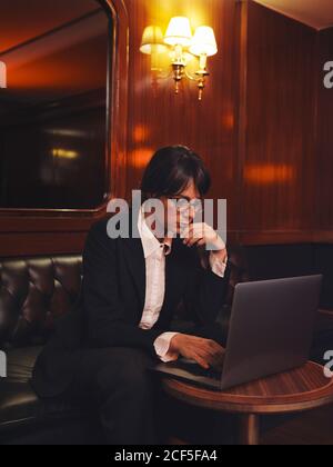 Femme d'affaires intelligente en lunettes dactylographiant sur ordinateur portable confortablement assis dans un canapé en cuir noir dans le café Banque D'Images