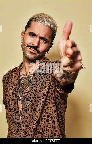 Homme barbu adulte avec une coupe de cheveux élégante et tatouage habillé chemise leopard faisant un geste de pistolet de doigt et regardant l'appareil photo sur fond jaune Banque D'Images