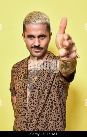 Homme barbu adulte avec une coupe de cheveux élégante et tatouage habillé chemise leopard faisant un geste de pistolet de doigt et regardant l'appareil photo sur fond jaune Banque D'Images