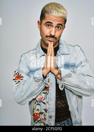 Jeune homme tendance dans une veste en denim élégante qui supplie avec des plis les mains près du menton et regardant l'appareil photo sur fond gris Banque D'Images