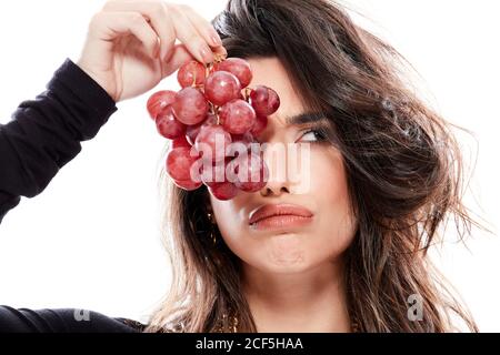 Magnifique femme à cheveux noirs dans des vêtements tendance tenant la branche de les raisins rouges devant la moitié de la face et en regardant loin faire des visages amusants sur un fond blanc isolé Banque D'Images