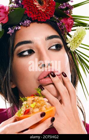 Jeune femme magnifique avec couronne de fleurs colorées sur la tête et le doigt sur la lèvre regardant loin tout en tenant le plat dans le zeste d'orange isolé sur fond blanc Banque D'Images