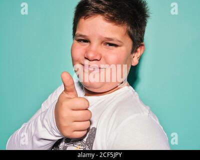 Joyeux et confiant Chubby grand garçon de la taille de la preteen dans des vêtements décontractés regarder l'appareil photo et rire tout en faisant des gestes contre fond turquoise Banque D'Images