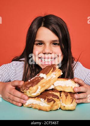 Gaie petite fille en tenue décontractée appréciant doux eclairs avec chocolat regardant l'appareil photo tout en étant assis à la table contre le rouge arrière-plan Banque D'Images