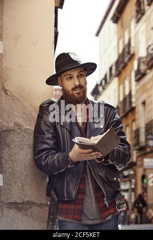 Jeune homme beau barbu en chapeau noir et veste en cuir avec livre ouvert dans les mains debout près du mur à l'intérieur rue et regarder le long Banque D'Images
