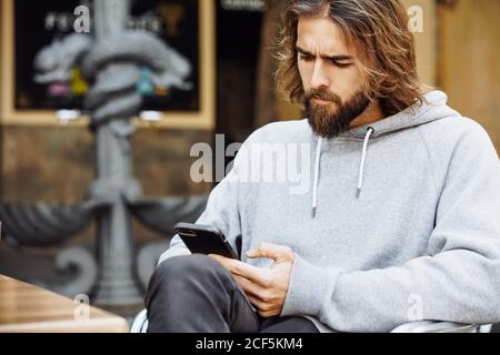 Jeune homme beau barbu dans un chandail décontracté assis au café concentré sur le téléphone mobile Banque D'Images
