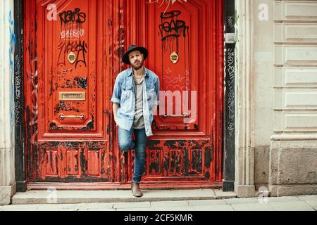Jeune homme beau barbu en chapeau et chemise en denim à la recherche le long du pied debout dans les portes peintes en rouge Banque D'Images